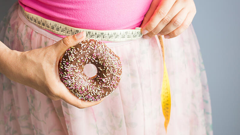 Woman holding donut in hand and check out his body fat with measuring tape.