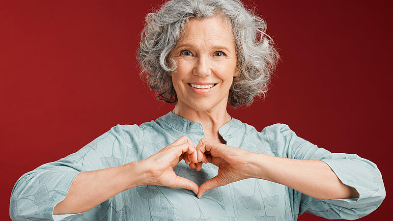 Love, heart and health retired woman with loving emoji sign, icon or symbol showing care or affection on red valentines day studio background. Grey, senior or pensioner with a trendy emoticon pose