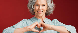 Love, heart and health retired woman with loving emoji sign, icon or symbol showing care or affection on red valentines day studio background. Grey, senior or pensioner with a trendy emoticon pose