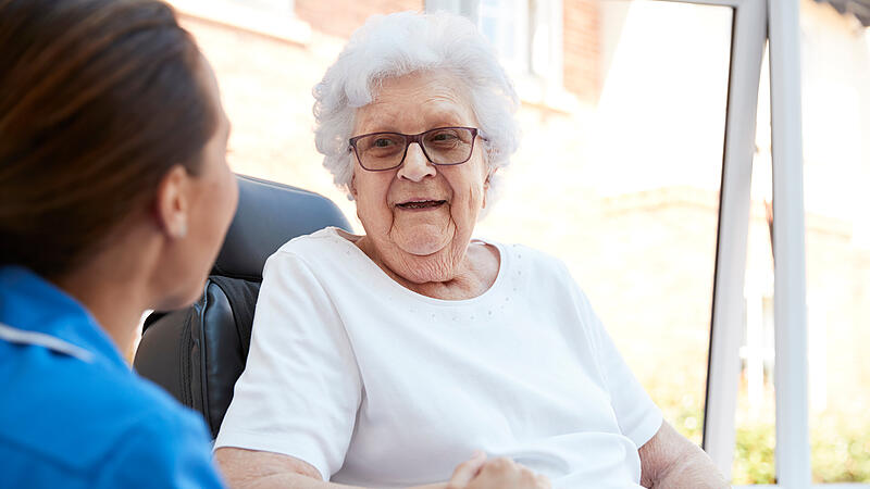 Senior Woman Sitting In Motorized Wheelchair Talking With Nurse In Retirement Home