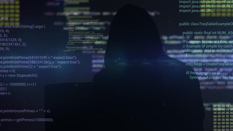 Unrecognizable man hacker wearing sweatshirt with hood typing on keyboard. Programming code on a large screen in the background.