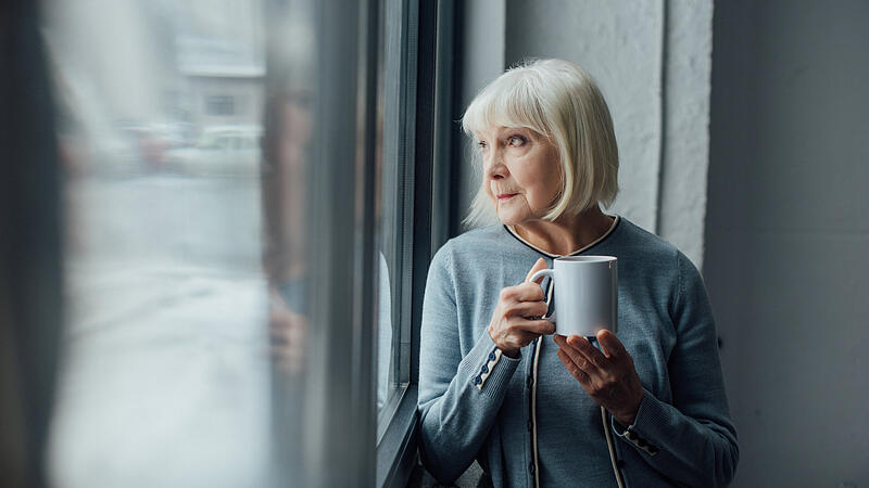 pensive senior woman holding cup of coffee and looking through window at home