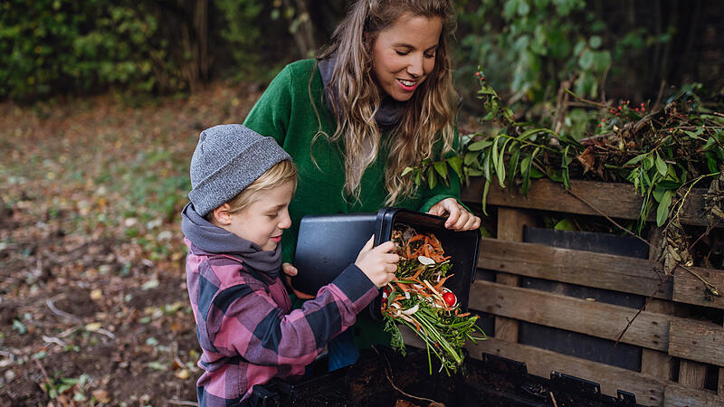 Boy helping mother to put kitchen waste, peel and leftover vegetables scraps into composter in the garden. Concept of composting kitchen biodegradable waste.